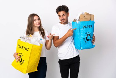 Beyaz arka planda izole edilmiş plastik ve kâğıt dolu bir çanta tutan genç çift, parmak kenarı işaret ediyor.