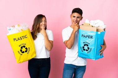 Pembe arka planda izole edilmiş plastik ve kağıt dolu bir çanta taşıyan genç çift yan tarafa bakıyor ve gülümsüyor.