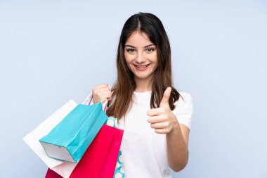 Genç esmer kadın izole edilmiş mavi arka planda elinde alışveriş çantaları ve başparmağıyla