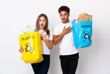 Beyaz arka planda izole edilmiş plastik ve kağıt dolu bir çanta taşıyan genç çift sağa bakarken şaşırmış ve şok olmuştu.