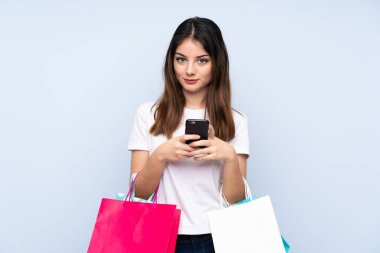 Genç esmer kadın izole edilmiş mavi arka planda elinde alışveriş torbalarıyla bir arkadaşına cep telefonuyla bir mesaj yazıyor.