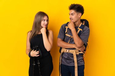 Sarı arka planda izole edilmiş büyük sırt çantalı genç dağcı çift birbirlerine bakıyor.