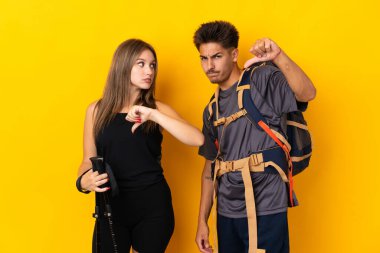 Sarı arka planda izole edilmiş büyük sırt çantalı genç dağcı çift olumsuz ifade ile baş parmak aşağı işareti