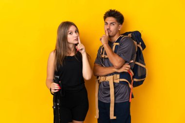 Sarı arka planda izole edilmiş büyük sırt çantalı genç dağcı çiftin ağzı kapanıyor ve sessizlik işareti var.