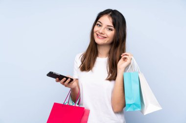 Genç esmer kadın izole edilmiş mavi arka planda elinde alışveriş torbalarıyla bir arkadaşına cep telefonuyla bir mesaj yazıyor.