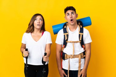 Sarı arka planda izole edilmiş büyük sırt çantalı genç dağcı çift yukarı bakıyor ve şaşırmış bir ifadeyle