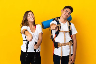 Sarı arka planda izole edilmiş büyük sırt çantalı genç dağcı çift mutlu bir ifadeyle önü işaret ediyor.