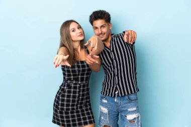 Mavi arka planda izole edilmiş genç beyaz çift gülümserken seni işaret ediyor.