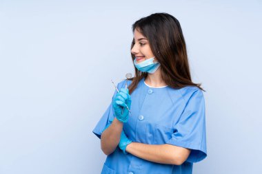 İzole edilmiş mavi arka planda aletleri tutan kadın dişçi yan tarafa bakıyor.