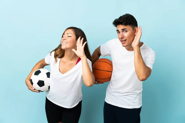 Young couple playing football and basketball isolated on blue background listening to something by putting hand on the ear