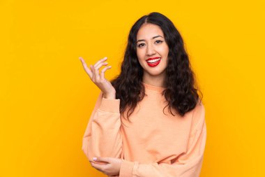 İspanyol Çinli kadın, izole edilmiş sarı arka planda. Davet edilmek için ellerini yana uzatıyor.