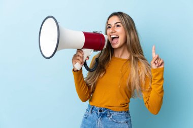 İzole edilmiş mavi arka planda genç İspanyol kadın megafondan bir şey duyurmak için bağırıyor.