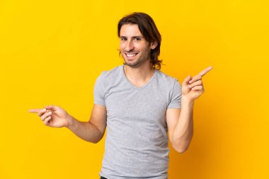 Sarı arka planda izole edilmiş genç yakışıklı adam parmak uçlarını işaret ediyor ve mutlu.