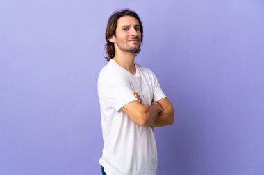 Mor arka planda soyutlanmış, kolları bağlı ve ileriye bakan genç yakışıklı bir adam.