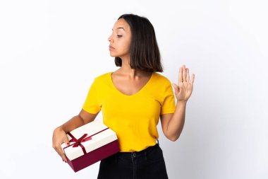 Beyaz arka planda izole edilmiş bir hediye tutan genç Latin kadın dur hareketi yapıyor ve hayal kırıklığına uğruyor.