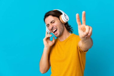 Genç yakışıklı adam mavi arka planda izole edilmiş müzik dinliyor ve şarkı söylüyor.
