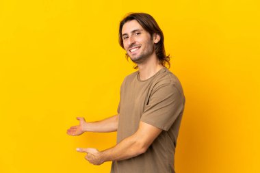Sarı arka planda izole edilmiş genç ve yakışıklı bir adam davet edildiği için ellerini yana uzatıyor.