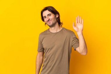 Sarı arka planda izole edilmiş genç yakışıklı adam mutlu bir ifadeyle selamlıyor.