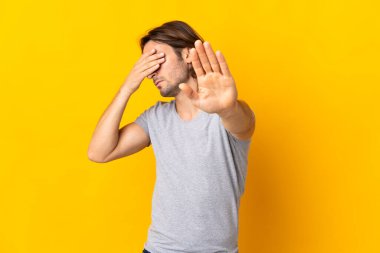 Sarı arka planda izole edilmiş genç yakışıklı adam dur hareketi yapıyor ve yüzünü kapatıyor.