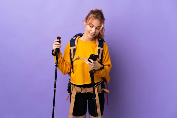 Teenager girl with backpack and trekking poles over isolated purple background sending a message with the mobile