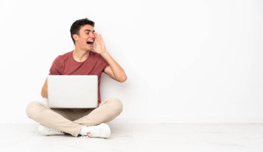 Genç adam diz üstü bilgisayarı ile salonda oturuyor ve ağzı sonuna kadar açık bağırıyor.
