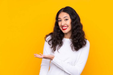 Karışık ırk kadını, izole edilmiş sarı arka planda. Davet etmek için ellerini yana uzatıyor.