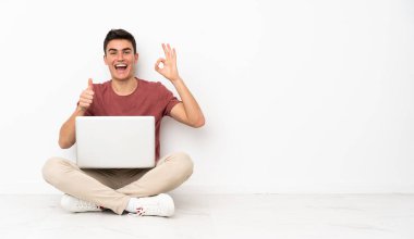 Genç adam fuayede dizüstü bilgisayarıyla oturuyor. Tamam işareti yapıyor ve el hareketi yapıyor.