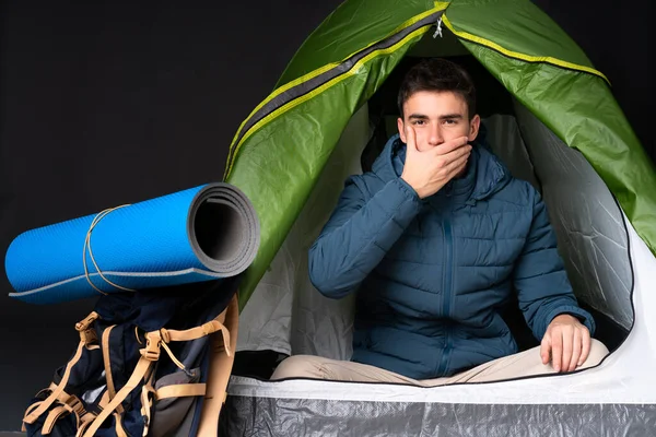 Teenager caucasian man inside a camping green tent isolated on black background covering mouth with hands