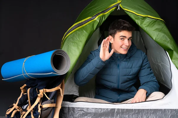 Teenager caucasian man inside a camping green tent isolated on black background listening to something by putting hand on the ear