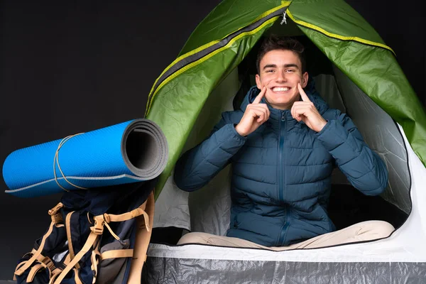 Teenager caucasian man inside a camping green tent isolated on black background smiling with a happy and pleasant expression