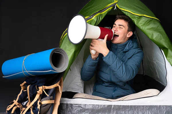 Teenager caucasian man inside a camping green tent isolated on black background shouting through a megaphone