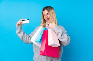 Genç Uruguaylı kadın izole edilmiş mavi arka planda elinde alışveriş çantaları ve kredi kartı tutuyor.