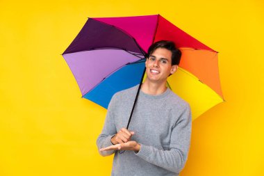 İzole edilmiş sarı arka planda şemsiye tutan adam, davet edilmek için ellerini yana uzatıyor.