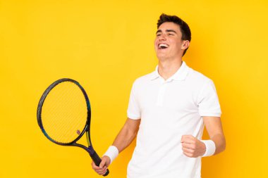 Yakışıklı genç tenisçi sarı arka planda izole edilmiş tenis oynuyor ve zaferi kutluyor.