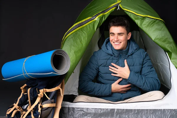Teenager caucasian man inside a camping green tent isolated on black background smiling a lot