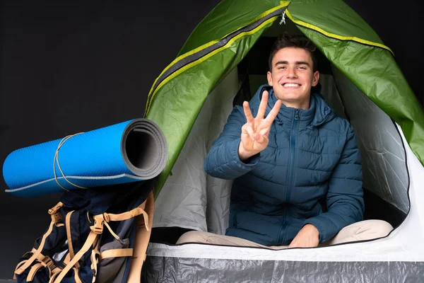 Teenager caucasian man inside a camping green tent isolated on black background happy and counting three with fingers