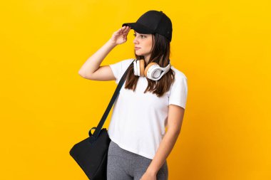 Spor çantalı genç bayan, sarı arka planda yalıtılmış, sürpriz bir ifadeyle yan tarafa bakarken.