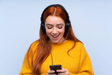 Kızıl saçlı genç kız mavi arka planda müzik dinliyor ve cep telefonu arıyor.