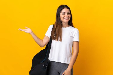 Spor çantalı genç bayan sarı arka planda izole edilmiş. Davet etmek için ellerini yana uzatıyor.