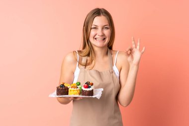 Ukraynalı aşçı pembe arka planda izole edilmiş bir kek tutuyor ve parmaklarıyla 