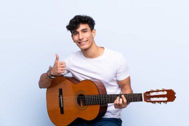 Gitarlı genç Arjantinli adam mavi arka planda, iyi bir şey olduğu için başparmak kaldırıyor.