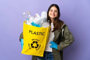 Geri dönüşüm için plastik şişelerle dolu bir çanta taşıyan genç bir kadın mor arka planda izole edilmiş, kolları kalçasında poz veriyor ve gülümsüyor.