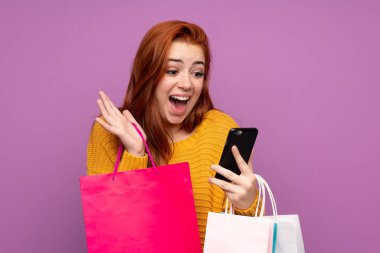 Mor arka planda kalan kızıl saçlı genç kız elinde alışveriş torbalarıyla bir arkadaşına cep telefonuyla bir mesaj yazıyor.