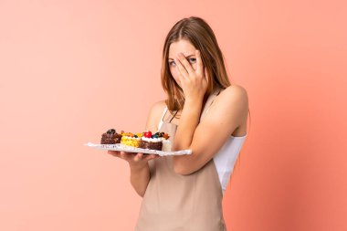 Ukraynalı pasta şefi pembe arka planda izole edilmiş bir kek tutuyor gözleri kapatıyor ve parmaklarından bakıyor.