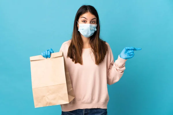 Young woman protecting from the coronavirus with a mask and holding a grocery shopping bag isolated on blue background surprised and pointing side
