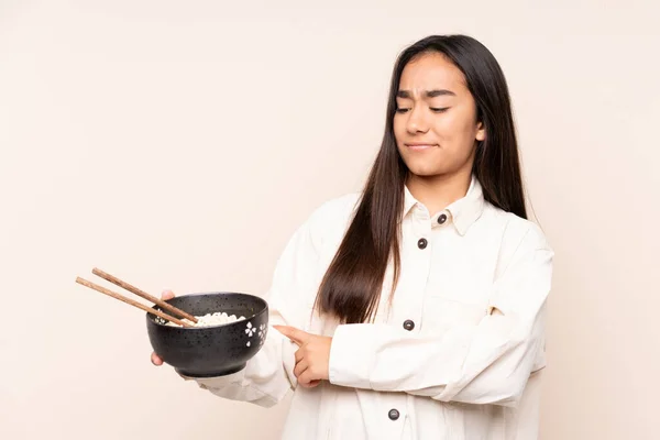 Young Indian woman isolated on beige background with sad expression while holding a bowl of noodles with chopsticks