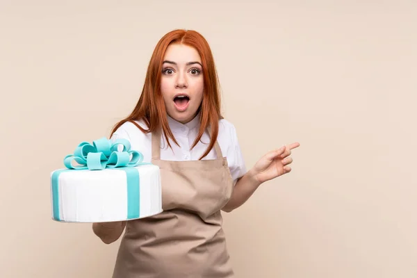 Redhead teenager girl with a big cake over isolated background surprised and pointing finger to the side