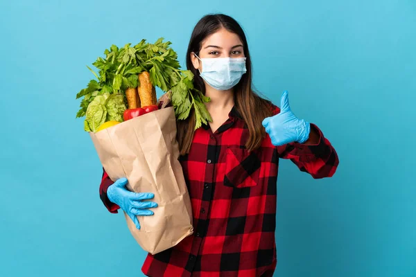 Young caucasian with vegetables and mask isolated on blue background with thumbs up because something good has happened