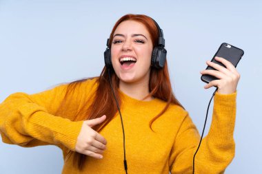 Kızıl saçlı genç kız mavi arka planda müzik dinliyor ve gitar jesti yapıyor.