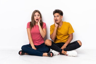Yerde oturan genç çift, beyaz arka planda izole edilmiş, şaşırmış ve şok olmuş bir yüz ifadesiyle.